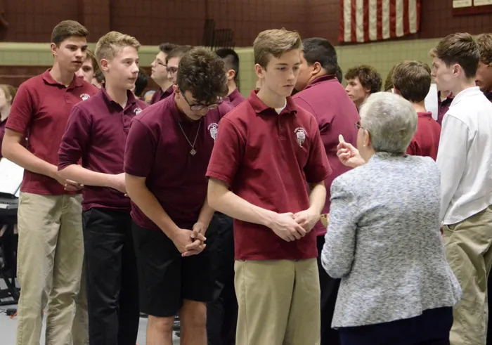 Young men in maroon shirts listening attentively to an older woman indoors.